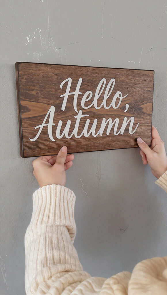 A person hanging a 'Hello, Autumn' wooden sign as an example of affordable autumn wall decor.
